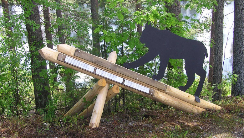 A metal, life-size statue of a lynx on a wooden pedestal at the edge of a forest. A lake is in the background.